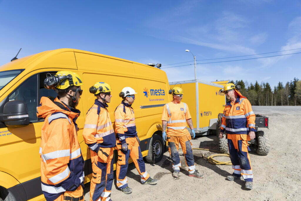 Fem arbeidere i reflektivt arbeidstøy som står foran en oransje arbeidsbil og prater med hverandre på en solskinsdag.